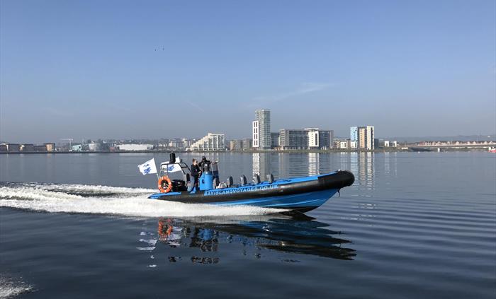 Blue Rib boat crossing Cardiff Bay with trailing wake behind it.  View of buildings in the background.